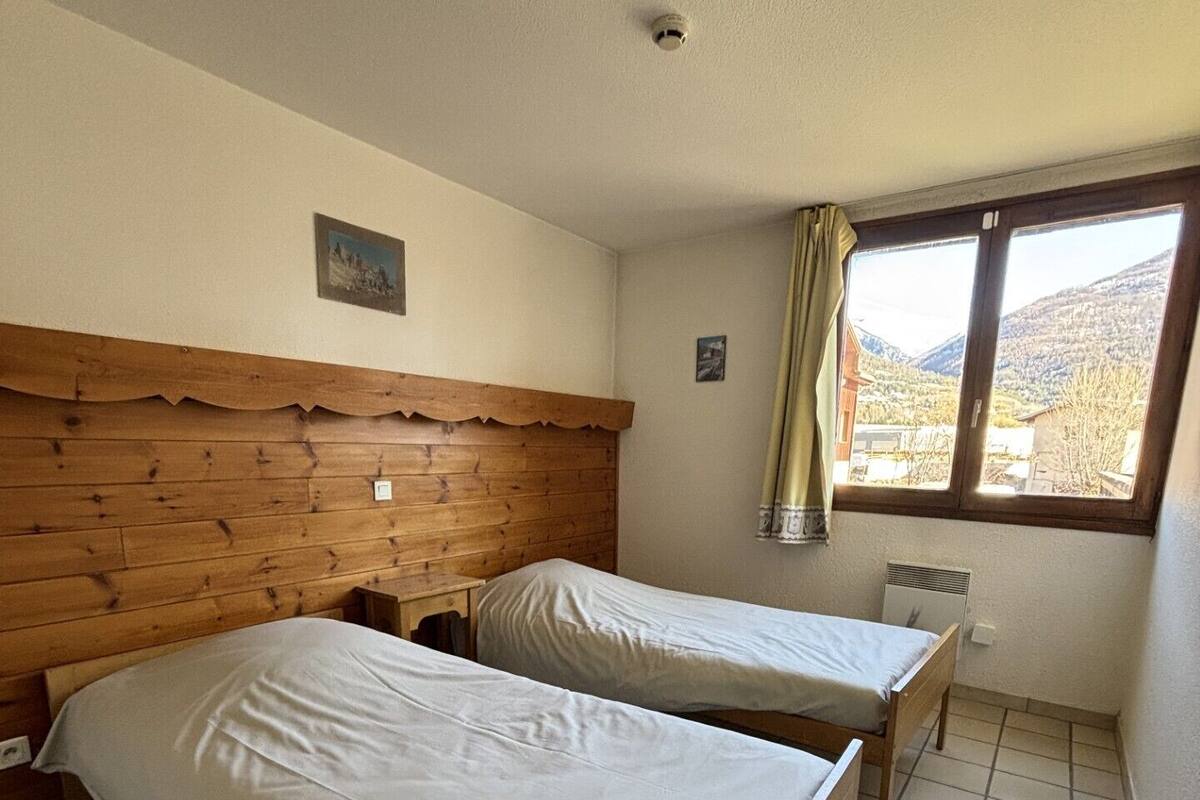 Une pièce avec deux lits en bois, une petite table, une fenêtre donnant sur les montagnes et un luminaire au plafond.
