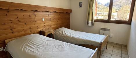 A room with two wooden beds, a small table, a window with a view of mountains, and a ceiling light.