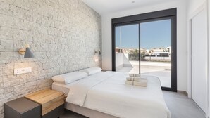 Une chambre moderne avec un mur en pierre, un grand lit, une table de chevet, une lampe et une vue sur l’extérieur, visible à travers une porte vitrée.