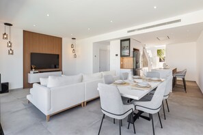Una sala de estar moderna con un sofá blanco, una mesa de comedor con sillas, un televisor de pantalla plana y lámparas colgantes.