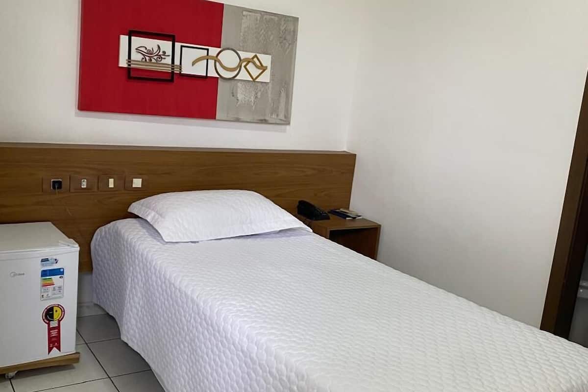 A single bed with a wooden headboard, a white quilted bedspread, a red and gray abstract artwork above the bed, and a small bedside table with a phone and a book.