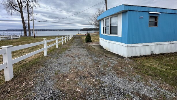A gravel path leading to a blue building with a white fence on one side and a view of a body of water in the background. - 2-bedroom mobile home in Henderson (Henderson)