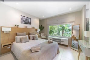 A modern bedroom with a large bed, bedside tables, a desk, a mirror, and a window with a view of greenery.