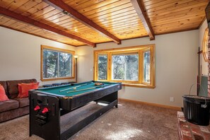 A pool table in a room with a wooden ceiling, a couch, and windows with natural light.