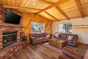 A cozy living room with a brick fireplace, wooden ceiling beams, and large windows.