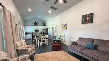 Una sala moderna con ventilador de techo, un sofá, sillones estampados, una zona de comedor y una cocina al fondo.