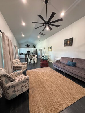 A modern living room with a ceiling fan, a sofa, patterned armchairs, a dining area, and a kitchen in the background. - Country Retreat + Game Room (McKinney)