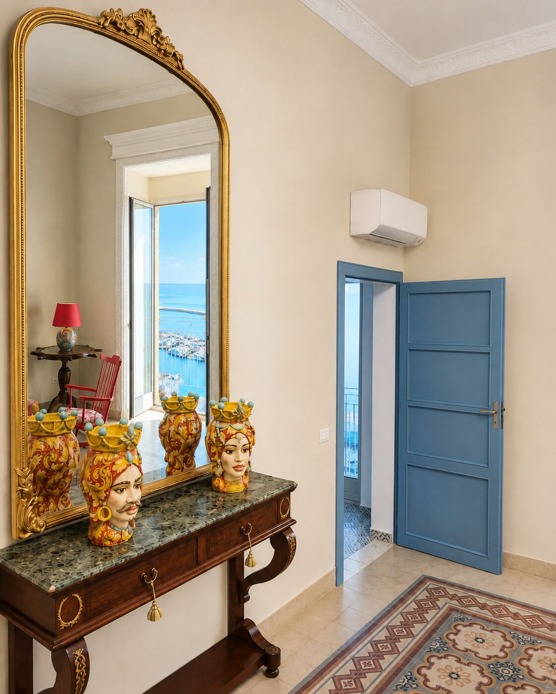 Une pièce dotée d’un grand miroir décoratif, d’une porte bleue et offrant une vue sur la mer par une fenêtre.