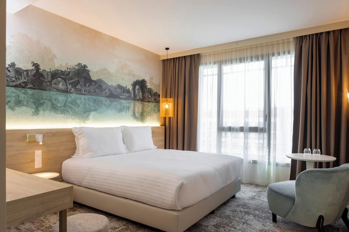 A hotel room with a large bed, a desk, a chair, a small table, a painting on the wall, and a view through the window.