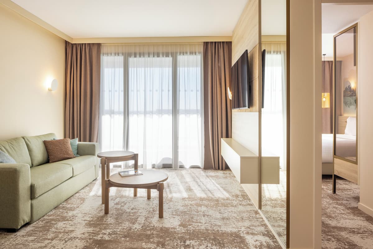 A hotel room with a sofa, a small round table, a mirror, and a large window with curtains.