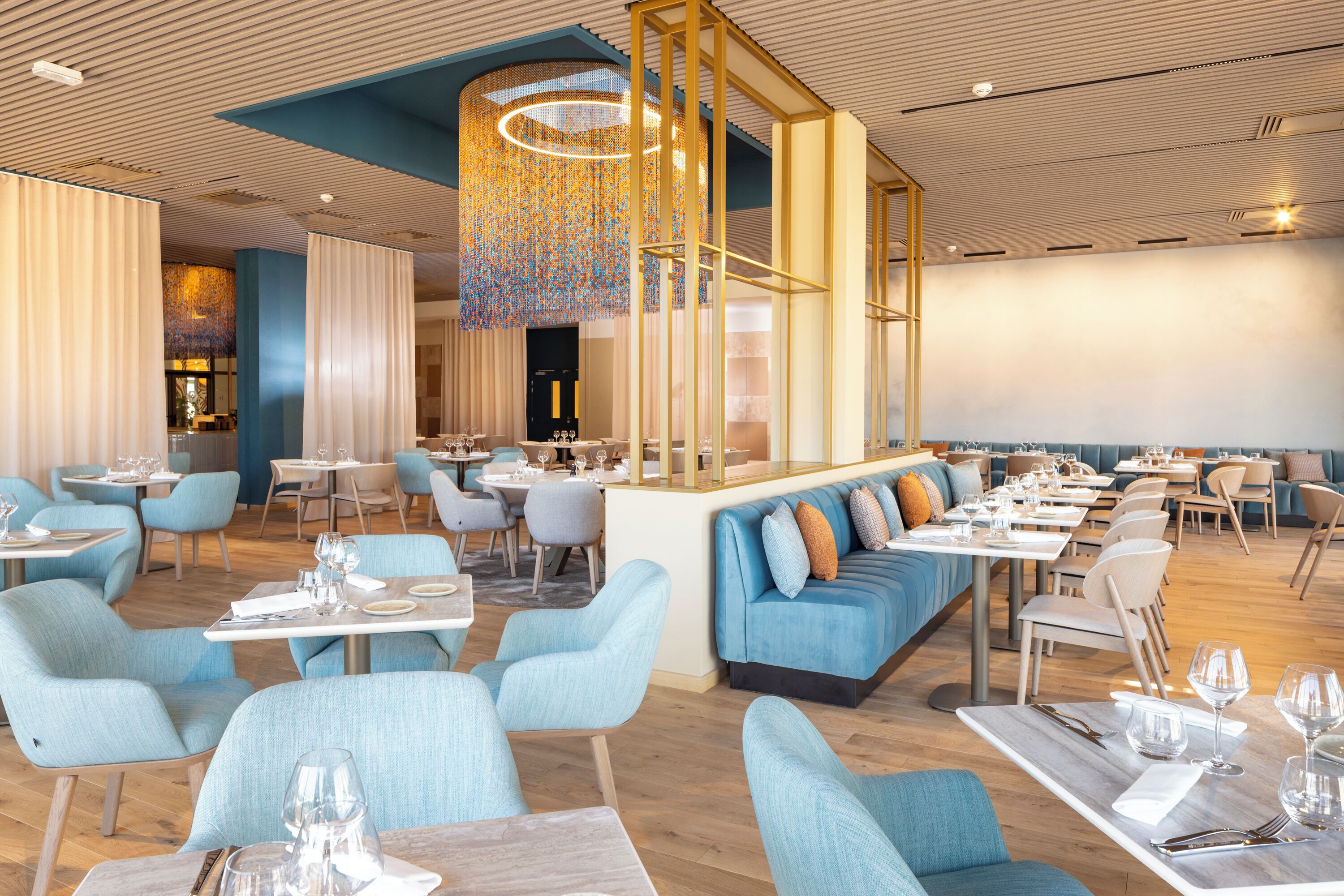 A modern restaurant interior with blue upholstered seating, wooden tables, and a central decorative light fixture.