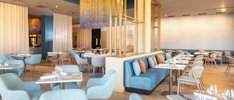A modern restaurant interior with blue upholstered seating, wooden tables, and a central decorative light fixture.