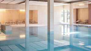 A modern indoor swimming pool with clear glass walls, lounge chairs, and a reception area. - Parkest Hôtel & Spa (Genas)