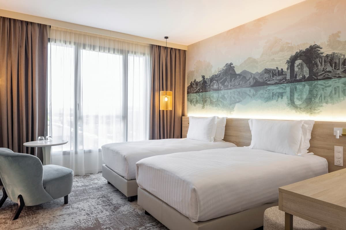 A hotel room with two beds, a small table, a chair, and a painting on the wall.