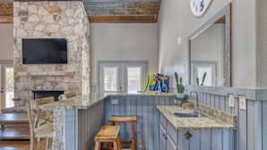 Una cocina con pared de piedra, taburetes de madera, un lavabo y un televisor sobre la encimera.