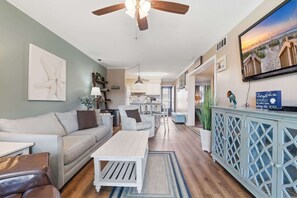 Un salon avec un canapé, une table basse et un téléviseur. On y trouve un ventilateur de plafond, une cuisine à l’arrière-plan et un meuble orné d’un objet décoratif.