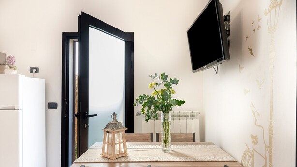A dining area with a table, chairs, a vase with flowers, a lantern, a wall-mounted TV, and a door. - Apartment 'Lu Marchese' with Private Terrace, Wi-Fi and Air Conditioning (Minervino di Lecce)