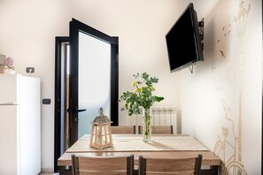A dining area with a table, chairs, a vase with flowers, a lantern, a wall-mounted TV, and a door.