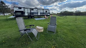 A grassy field with a trailer, two lawn chairs, a hammock, and a picnic table. - Haven on Wheels- Cozy Travel Trailer Getaway in Nature (Williamston)