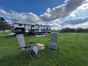 Ein grasbewachsenes Feld mit einem Wohnwagen, zwei Liegestühlen, einer Hängematte und einem Picknicktisch.
