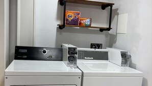 A laundry area with two white washing machines, a shelf with detergent boxes, and a wall-mounted light switch. - Cozy Albert St Retreat (Waterloo)