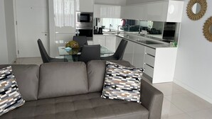 A modern kitchen with white cabinets, a dining area with a glass table and chairs, and a gray sofa with patterned cushions.