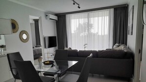 A modern living room with a glass dining table, black chairs, a dark sofa, a flat-screen TV, and a large window with curtains.