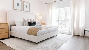 A modern bedroom with a bed, bedside tables, a lamp, and a large window with curtains. - Luxury 4-bed, 3.5-bath home in Shelter Bay, West K,  rooftop deck with hot tub. (West Kelowna)
