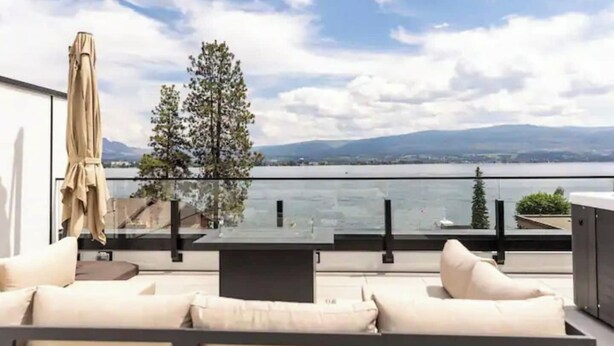 A balcony with a sofa, a table, and an umbrella, overlooking a lake and mountains. - Luxury 4-bed, 3.5-bath home in Shelter Bay, West K,  rooftop deck with hot tub. (West Kelowna)