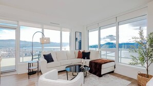 A modern living room with a white sofa, a round coffee table, and a view of a cityscape through large windows. - Skyfall 3 Bed 1 Den Lakeview Amenity Rich 30 Floor (Kelowna)