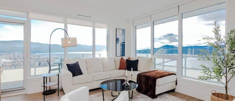 Una sala de estar moderna con un sofá blanco, una mesa de centro redonda y vistas a un paisaje urbano a través de amplios ventanales.