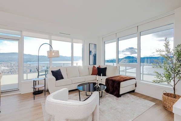 Ein modernes Wohnzimmer mit einem weißen Sofa, einem runden Couchtisch und Blick auf eine Stadtlandschaft durch große Fenster.
