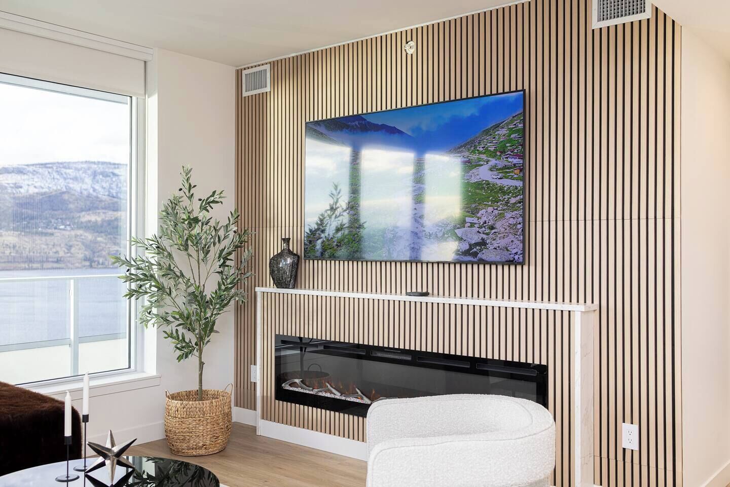 A modern living room with a fireplace, a large TV mounted on a wooden wall, a potted plant, and a view of a mountainous landscape through a large window.