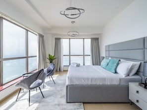 Un dormitorio moderno con una cama grande, una butaca tapizada de gris, una mesita de noche y vistas de la ciudad a través de amplias ventanas.
