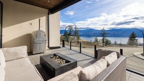 A modern outdoor seating area with a stone fire feature, a grill, and a view of a lake and mountains.