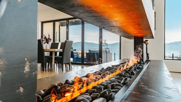 A long, narrow fire feature with rocks and flames, a modern interior with a dining area, and a view of the outdoors.