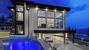 A modern two-story house with a swimming pool, a car parked outside, and a view of the landscape.