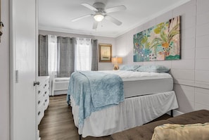 Ein Schlafzimmer mit Bett, Deckenventilator, einem Gemälde und einem Fenster mit Vorhängen.
