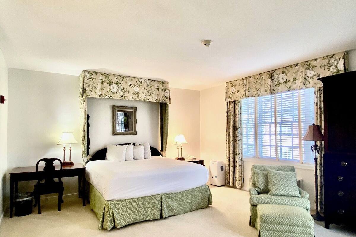Une chambre d’hôtel avec un grand lit, un bureau, une chaise, une commode et une fenêtre avec des rideaux.