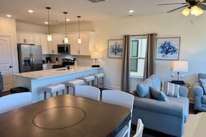 Une cuisine moderne avec des meubles blancs, un îlot central doté de tabourets de bar, une table à manger et un ventilateur de plafond.