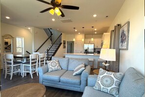 Un salon avec un canapé gris, des coussins à motifs, un ventilateur de plafond, un éclairage encastré, un escalier et un coin cuisine.