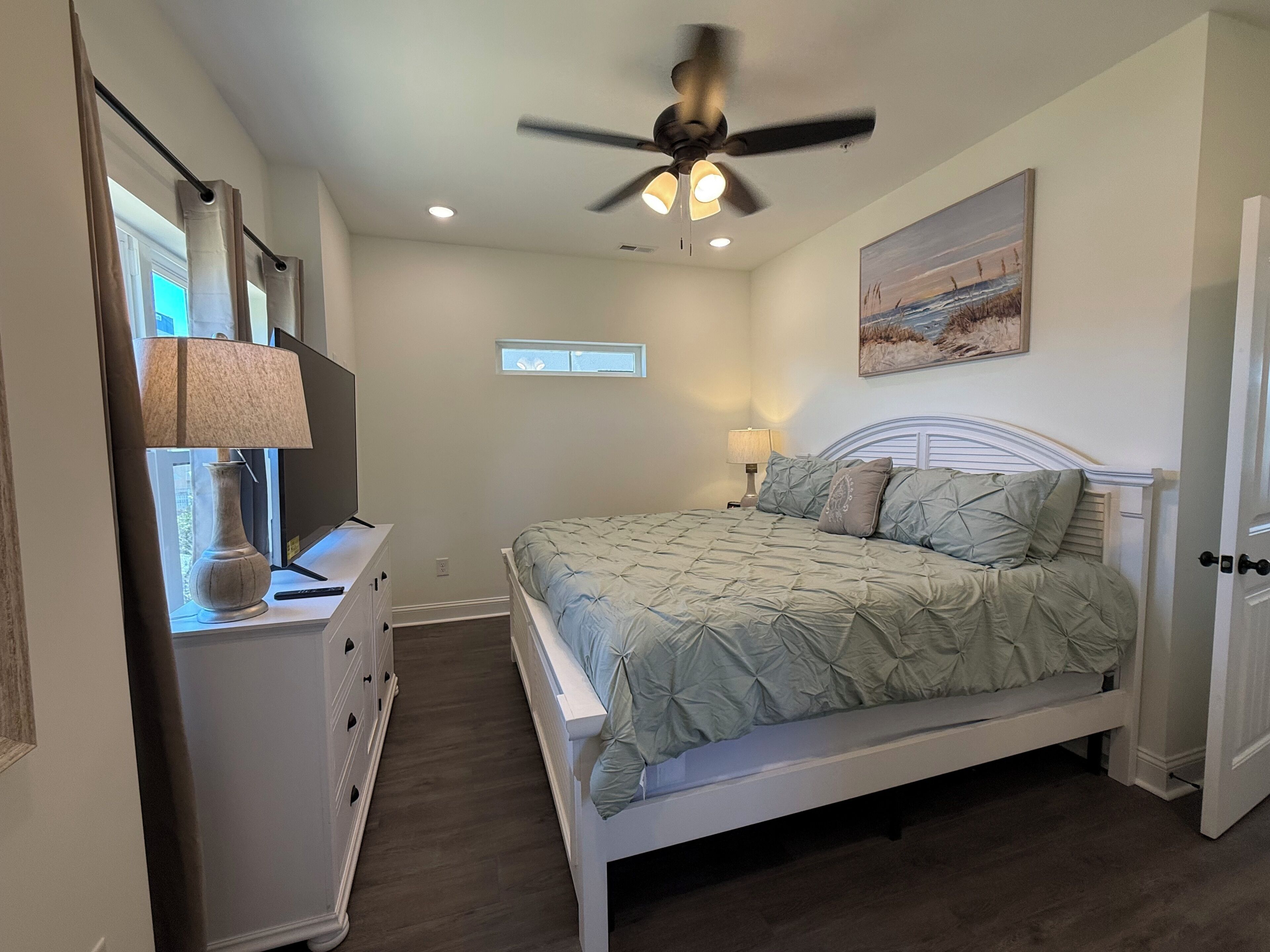 A bedroom with a bed, a ceiling fan, a window, a painting, and a dresser.