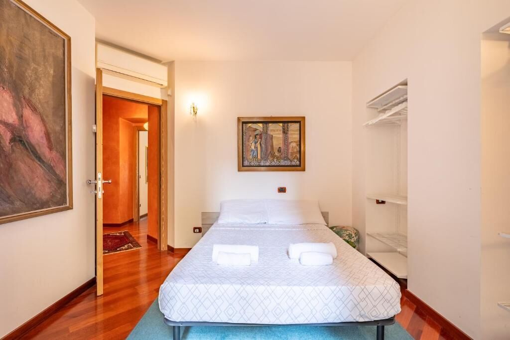 A single bed with a patterned bedspread, a painting on the wall, and a hallway leading to another room.