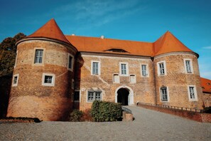 Un bâtiment historique en briques, doté d’une entrée centrale en arc de cercle, de deux tours rondes surmontées de toits coniques et d’une cour pavée.