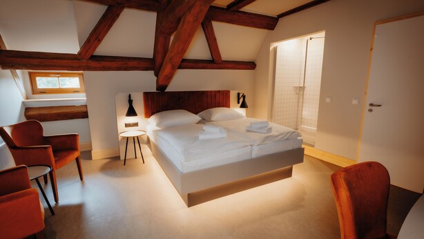 A bedroom with a large bed, wooden beams, a window, a chair, and a door. - Die Burg (Bad Belzig)
