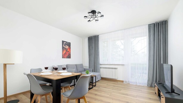 Ein modernes Wohnzimmer mit Esstisch, grauen Stühlen, einer Couch, einem Fernseher und einem großen Fenster mit Vorhängen.