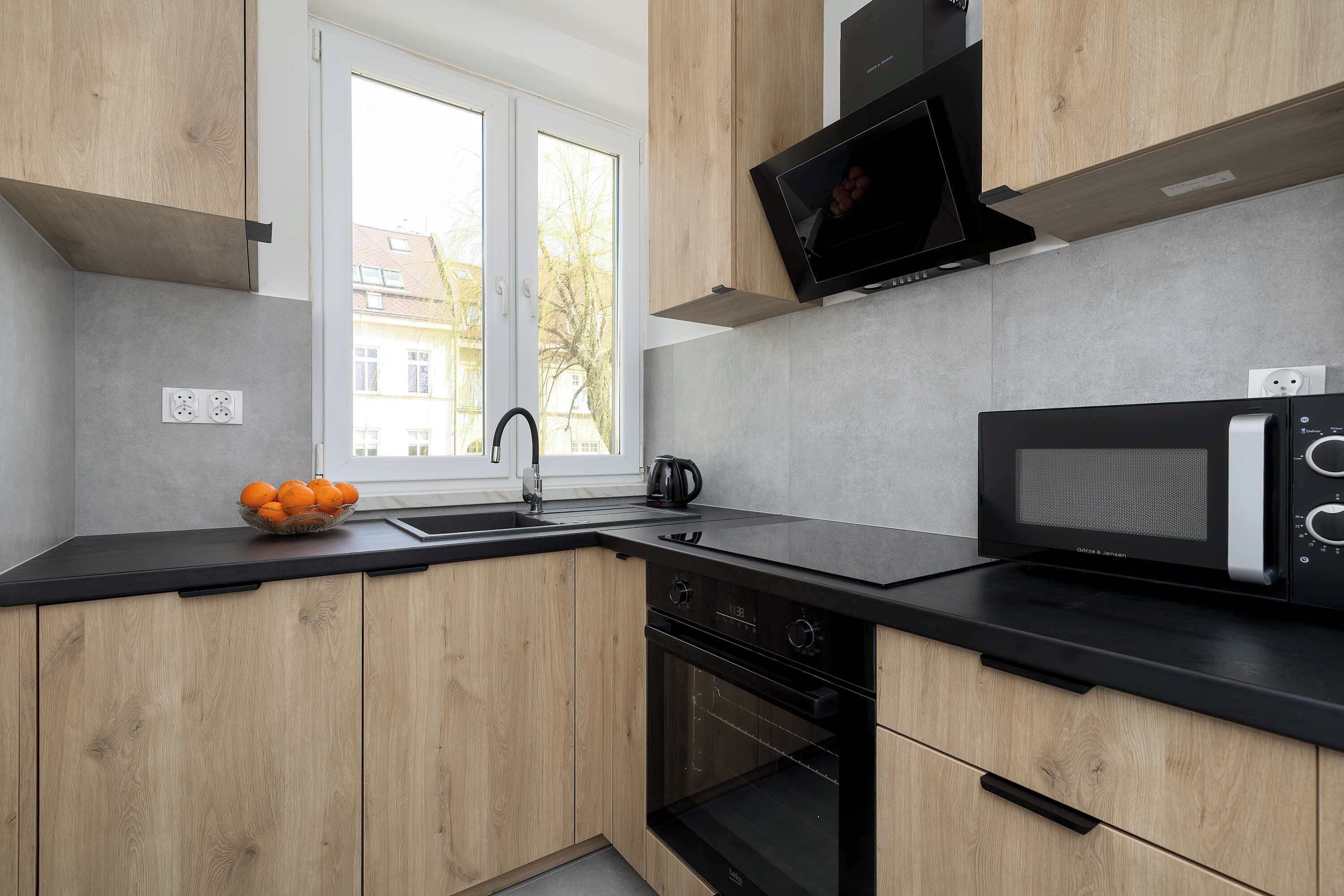 A modern kitchen with wooden cabinets, a black countertop, a built-in oven, a microwave, and a window with a view of buildings.