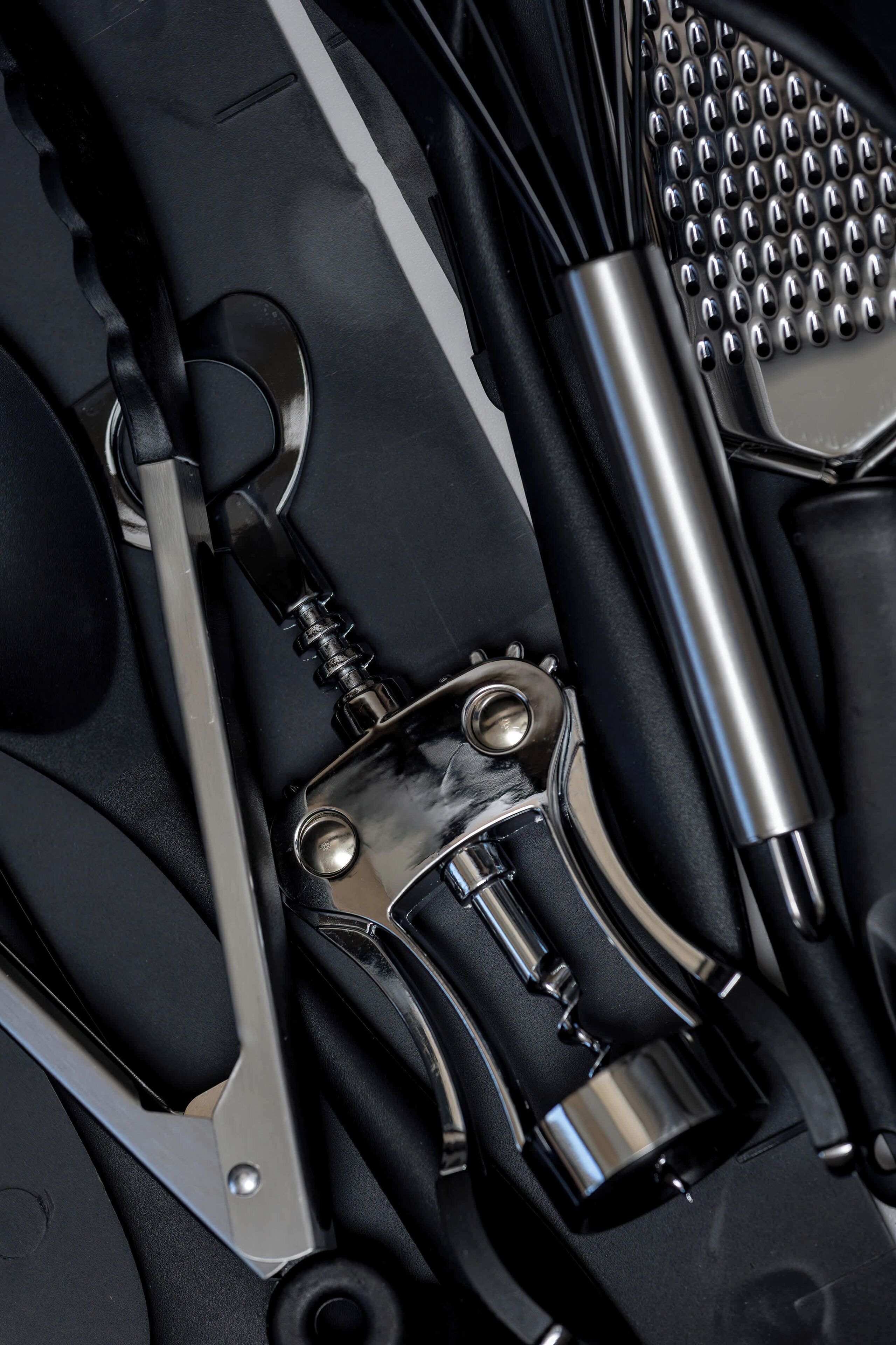 A collection of dark kitchen utensils and a corkscrew are arranged artistically in a drawer or tray. The overhead lighting creates sharp highlights on the metallic and black surfaces.