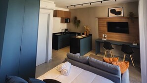 Een moderne hotelkamer met een bank, een bed met een opgerolde handdoek, een klein keukenhoekje, een bureau en een aan de muur bevestigde televisie.