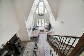 A high-ceilinged room with a staircase, a dining area with a table and chairs, and a seating area with sofas and a round table. - Lakeview Splendor- 7-BR Penthouse Bliss Rooftop (Harrison Hot Springs)
